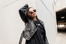 Urban beautiful young hipster woman with stylish round sunglasses in a fashion leather jacket with a black sweatshirt walks in the city