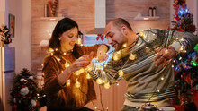 Cheerful Couple Getting Tangled In String Of Twinkle Lights For Christmas Eve Preparations. Man And Woman Laughing While Untangling Knot Of Garland With Lights And Illuminated Bulbs