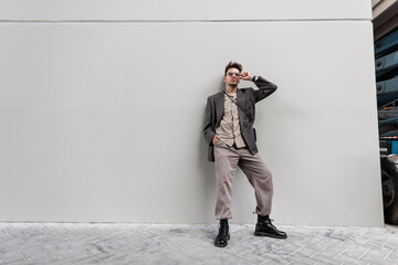 Fashionable young man in stylish casual look clothes with oversized blazer, shirt and black shoes stands near a gray wall in the city