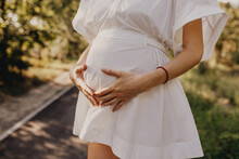 Closeup of a pregnant woman in white dress, holding hands on belly, outdoors.