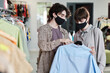 © AnnaStills - Young woman in mask choosing a new shirt with the help of shop assistant in the shopping mall