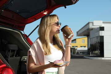 Beautiful young woman with doughnut drinking coffee near car at gas station