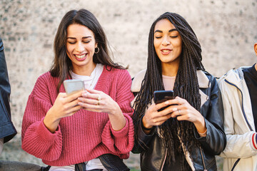 Two women best friends using smartphone together standing outdoors. Gen z young people using social network and new technology concept.