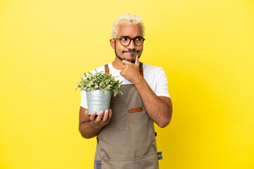 Wall Mural - Young Colombian man holding a plant isolated on yellow background having doubts and thinking