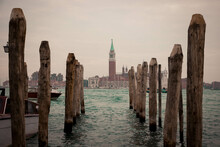Wooden Pillars In Water At A Venice Dock Symmetrically Aligned With An Island In Front On A Cloudy Day