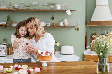 Happy chef cook baker mom woman in white shirt work with baby girl helper use hold mobile cell phone smile at kitchen table home Cooking food process concept Mommy little kid daughter prepare cake