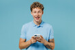 Surprised shocked vivid satisfied smiling overjoyed young curly man 20s years old wears azure t-shirt hold in hand use mobile cell phone isolated on plain pastel light blue background studio portrait