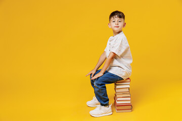 Side view young smart wunderkind school boy 5-6 years old in t-shirt casual clothes sit on pile of books isolated on plain yellow background studio Childhood children kids education lifestyle concept.