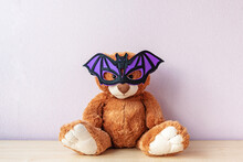 Funny Brown Teddy Bear With Purple Bat Shaped Mask Sits On Wooden Table By Light Pink Wall In Room. Celebrating Halloween
