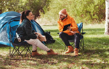 Poster - camping, tourism and travel concept - happy couple with tablet pc computer and book resting at tent camp