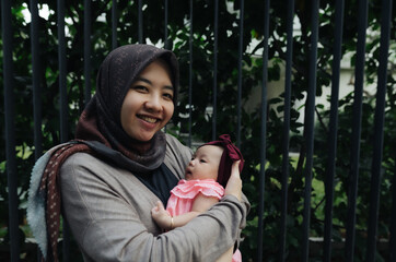 Wall Mural - Happy asian mom holding her little baby daughter wearing hijab