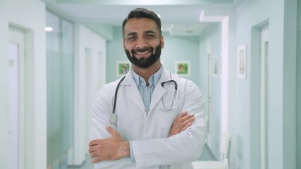 Wall Mural - Smiling happy cheerful male Indian latin doctor medical worker in white robe with stethoscope around neck standing in modern hospital clinic with arms crossed looking at camera. Headshot portrait.