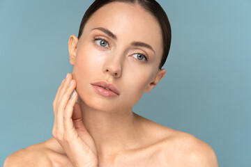 Studio portrait of young girl with natural makeup, combed hair, touching well-groomed pure skin on face, isolated on blue background. Beauty care, facelift, anti-aging cosmetic procedure concept