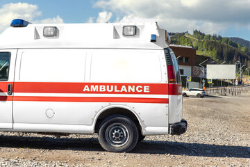 Side view of white ambulance rescue ems van car parked near countryside rural road at highland mountain resort area. Paramedic first aid help service vehicle against alpine forest landscape