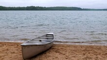 An Aluminum Canoe Is Beached On The Sand Of A Minnesota Lake Beach As Gentle Waves Roll In To Shore, In Early Morning Light On A Cloudy Day.