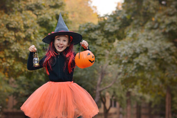 Wall Mural - Halloween trick or treat!A happy girl in a carnival witch costume holds a bucket in the form of a pumpkin for sweets and a lantern on the background of an autumn park.Happy Halloween!