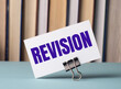 A white card with the text REVISION stands on a clip for papers on the table against the background of books. Defocus
