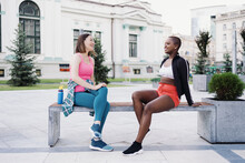 Cheerful Smiling Friends In Sportswear Sitting On Bench In The City Dicussing In Park. Multiethnic Women Having A Fitness Workout Break.