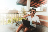 Fototapeta  - Mężczyzna, cyfrowy nomada pracujący z laptopem i rozmawiający przez telefon komórkowy w słoneczny dzień.