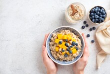 Oatmeal Bowl With Fruits And Berries In A Young Woman's Hands. Cooked Oats Porridge With Mango And Blueberries Over Grey Concrete Background, Table Top View, Copy Space