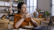 © PRPicturesProduction - portrait pretty asian businesswoman sitting at dining table behind computer is holding morning coffee and gazing into distance in a good mood.
