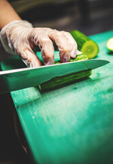 Wall Mural - woman hands in disposable gloves slicing cucumber on cutting board.