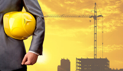 engineer holding helmet for workers security on background of new buildings and construction cranes on background of evening sunset sky. Silhouette building constructions with crane lifts load