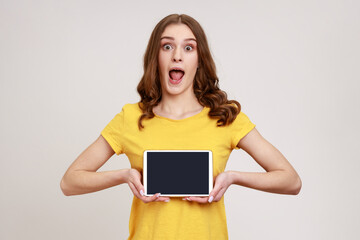 Wall Mural - Portrait of shocked beautiful teenage girl in style T-shirt, showing tablet empty screen with unbelievable face, looking at camera with opened mouth. Indoor studio shot isolated on gray background.