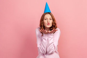 Poster - Portrait of happy curly haired teenage girl in hoodie with party hat sending air kiss at camera, holiday greeting, flirt. Indoor studio shot isolated on pink background