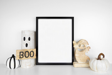 Wall Mural - Halloween mock up. Black frame on a white shelf with rustic wood ghost decor and pumpkins. Portrait frame against a white wall. Copy space.