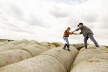 Brother Helping Sister Climbing On Rolled Hay Bales On Farm