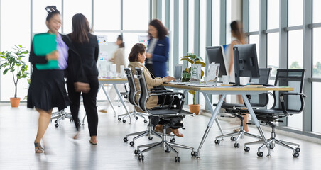 Wall Mural - Selective focus on one business woman sitting and working on laptops on conference table with other blurry business women walking in rush in office with multiple glass windows