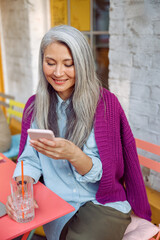 Wall Mural - Pretty senior Asian lady with grey hair reads sms on mobile phone holding drink at table on outdoors cafe terrace