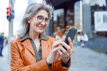 Positive hoary haired mature lady with glasses uses mobile phone standing on modern city street on autumn day