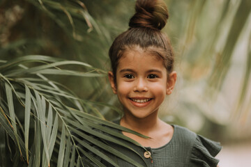 Wall Mural - The face of a little girl surrounded by tropical leaves. Closeup portrait of a beautiful swarthy baby with perfect skin and dark hair. Natural cosmetics, health, cleanliness, skin care, beauty concept