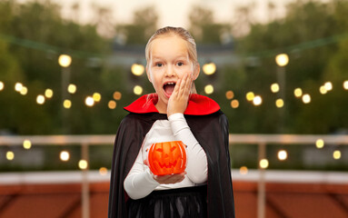 Wall Mural - halloween, holiday and childhood concept - happy girl in black dracula cape or costume with jack-o-lantern pumpkin over roof top party background