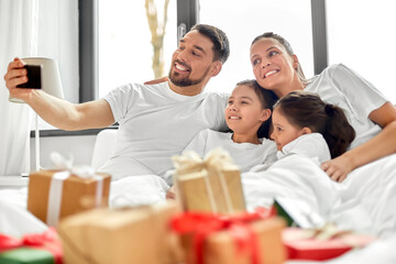 Poster - family, winter holidays and people concept - happy mother, father and two daughters with christmas gifts taking selfie in bed at home