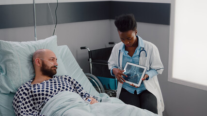 Wall Mural - Specialist surgeon black woman doctor explaining illness diagnosis showing x-ray using tablet computer in hospital ward. Sick man patient resting in bed discussing disease recovery treatment