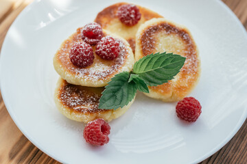 Delicious cheesecakes or pancakes with powdered sugar on white plate on wooden table with raspberries and mint leaf