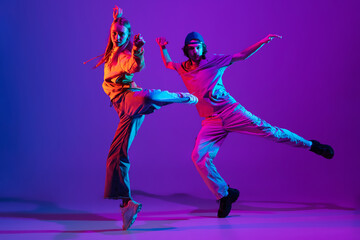 Wall Mural - Two dancers, young man and woman dancing hip-hop in casual sports youth clothes on gradient purple pink background at dance hall in neon light.