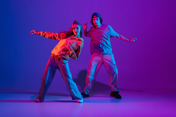 Wall Mural - Young man and woman dancing hip-hop in casual sports youth clothes on gradient purple pink background at dance hall in neon light.