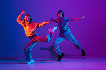 Wall Mural - Young man and woman dancing hip-hop in casual sports youth clothes on gradient purple pink background at dance hall in neon light.