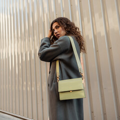 Wall Mural - Fashionable beautiful young curly woman in a stylish vintage coat with a handbag near a metal wall. Women's beauty and fashion