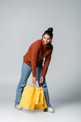 Wall Mural - full length of cheerful young woman in sweater and jeans holding yellow shopping bags on grey