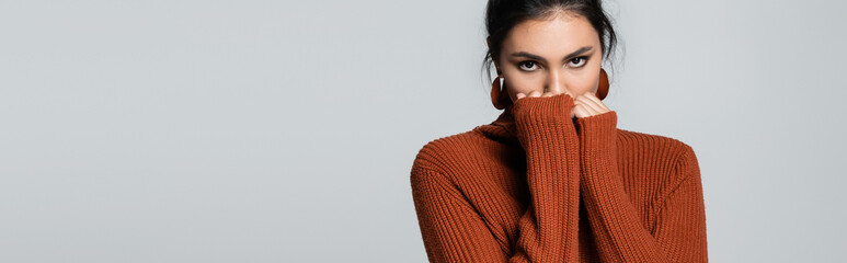 Wall Mural - young woman in knitted sweater looking at camera while covering face isolated on grey, banner