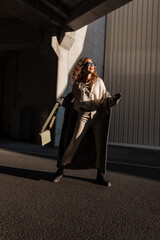 Wall Mural - Funny beautiful young smiling woman with curly hair in fashionable clothes look with a vintage coat, sunglasses, sweater, shoes and a handbag walks in the city on a sunny day. Happy girl with a smile