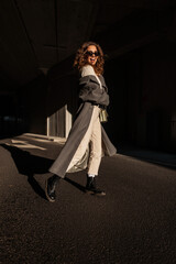 Wall Mural - Funny fashionable happy woman with curly hair and sunglasses in vintage long coat and stylish handbag walks in the city. Urban female fall style and happy emotions