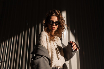 Wall Mural - Sunlight portrait of a beautiful young girl with curly hair with stylish sunglasses in a fashionable coat and sweater walks on the street near a metal wall. Female casual style and fashion