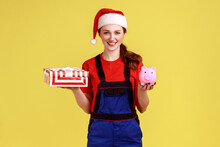 Happy Delivery Woman Holding Wrapped Gift Box And Piggy Bank, Savings For Online Delivering Service, Wearing Blue Overalls And Santa Claus Hat. Indoor Studio Shot Isolated On Yellow Background.