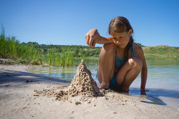 A little cute girl in a swimsuit is building a sand castle on the shore of a blue quarry lake.